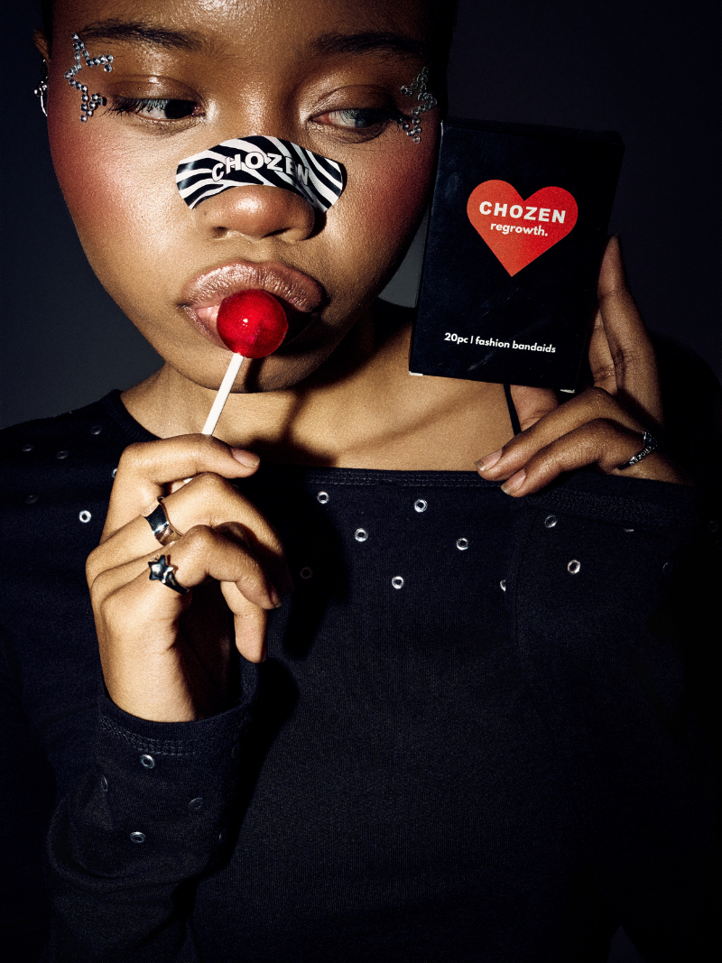A model wearing a black top with silver studs holds a black box labeled 'CHOZEN regrowth' next to a red lollipop. The model has silver glitter face decals and is wearing rings.